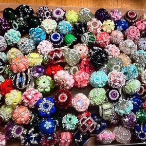 May include: A collection of colorful, decorative beads in various shapes and sizes. The beads feature floral and abstract designs, with a mix of colors including pink, blue, green, and purple. Some beads have pearl and rhinestone accents.