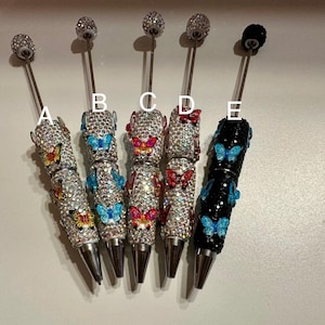 May include: Five decorative pens with rhinestone-covered barrels and butterfly embellishments. The pens feature silver-coloured barrels and different coloured butterfly accents, including yellow, blue, and red. Each pen has a different coloured top, including silver and black.