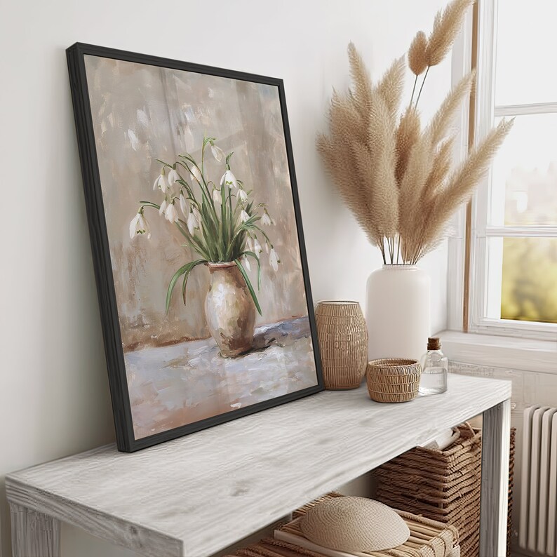 May include: A framed painting of a white flower bouquet in a brown vase on a light brown background. The painting is hanging on a white wall above a wooden shelf with a vase of dried pampas grass and other decorative items.