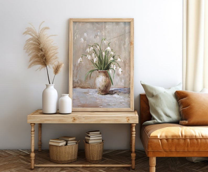 May include: A framed painting of a white flower bouquet in a brown vase on a white tablecloth. The painting is in a wooden frame and is hanging on a white wall.