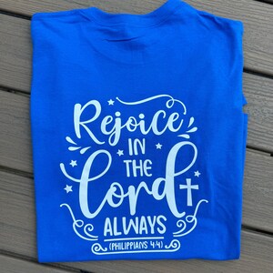 May include: A folded royal blue t-shirt with the words "Rejoice in the Lord Always" in white script. The text includes a cross and stars, with the scripture reference (Philippians 4:4) at the bottom.