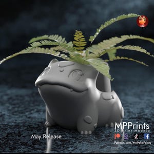 May include: A grey 3D-printed Bulbasaur planter holding green fern plants. The planter has a detailed design and the text "May Release" is visible. This unique planter is ideal for plant enthusiasts. The planter is a unique item for plant lovers.