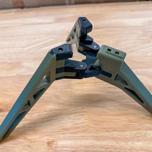 May include: A three-legged tripod stand featuring olive green and black components. The structure consists of three angled legs with triangular cutouts, connected by a central black hinge mechanism with visible silver screws at each joint.