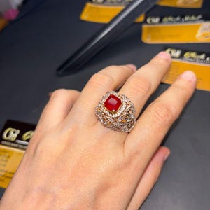 May include: A gold ring with a large, square, red gemstone surrounded by smaller, clear gemstones. The ring is on a person's finger.