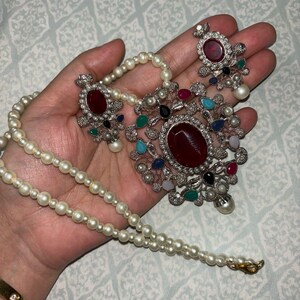 May include: A silver necklace with a large red gemstone pendant surrounded by smaller colorful gemstones. The necklace is strung with white pearls and has a matching pair of earrings with red gemstones.