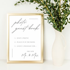 May include: A gold-framed sign with a white background and black text that reads "photo guest book". The sign has instructions for guests to take a photo, place it in the book, and leave a message for the new Mr. & Mrs.