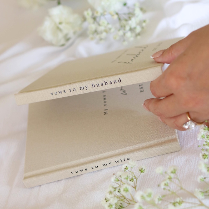 Peut inclure: Deux livres de v&oelig;ux beiges, dont un ouvert, avec le texte "vows to my husband" et "vows to my wife" imprim&eacute; sur les tranches. Le livre ouvert affiche du texte manuscrit. Le fond comprend des fleurs blanches et une main avec une bague.