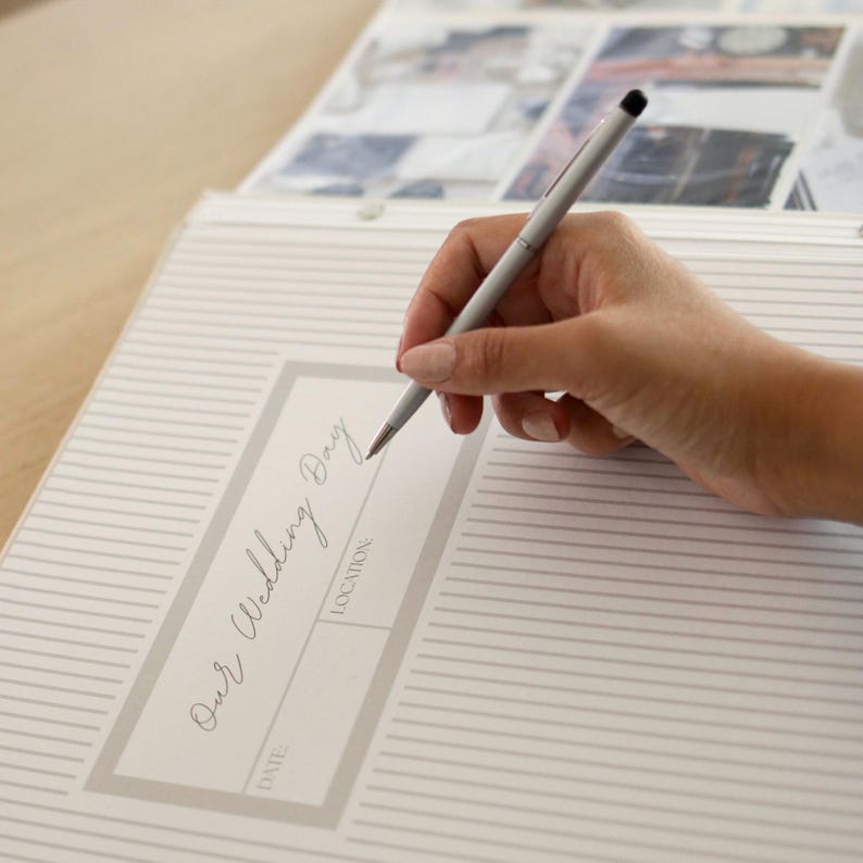 May include: A person writing in a wedding memory book with a silver pen. The book has a light gray striped pattern and the words "Our Wedding Day" are visible. The background shows other pages with photos.