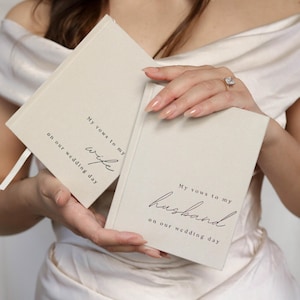 Peut inclure: Deux carnets de v&oelig;ux de mariage blanc cass&eacute; avec une &eacute;criture noire. L'un des livres se lit "Mes v&oelig;ux &agrave; ma femme le jour de notre mariage", et l'autre "Mes v&oelig;ux &agrave; mon mari le jour de notre mariage". Les livres sont tenus par une personne portant une robe blanche.