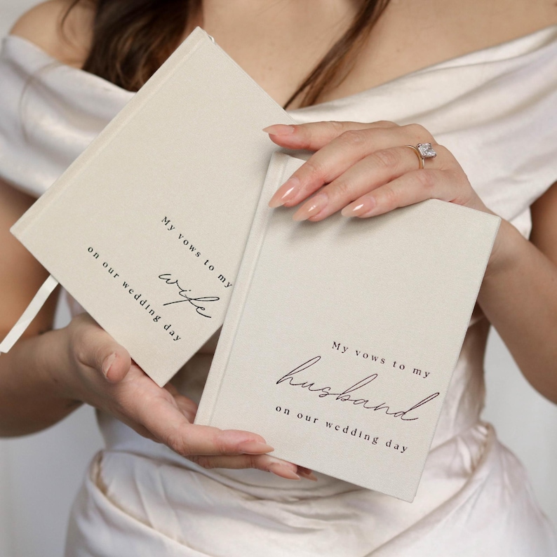 Peut inclure: Deux carnets de v&oelig;ux de mariage blanc cass&eacute; avec une &eacute;criture noire. L'un des livres se lit "Mes v&oelig;ux &agrave; ma femme le jour de notre mariage", et l'autre "Mes v&oelig;ux &agrave; mon mari le jour de notre mariage". Les livres sont tenus par une personne portant une robe blanche.