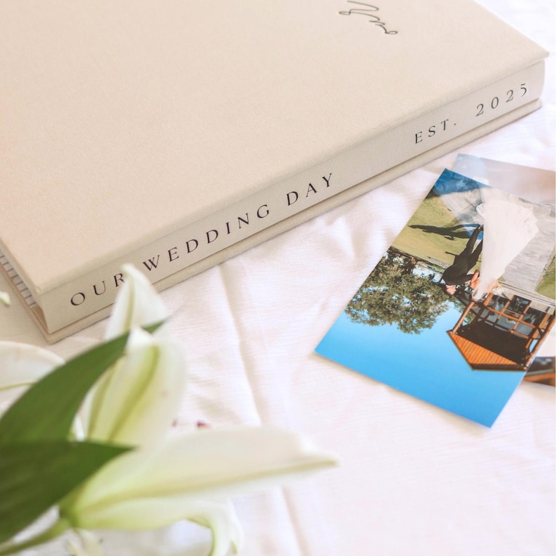 May include: A cream-colored wedding album with the text "OUR WEDDING DAY" and "EST. 2025" printed on the spine. A photograph is next to the album, and a white lily is in the foreground.