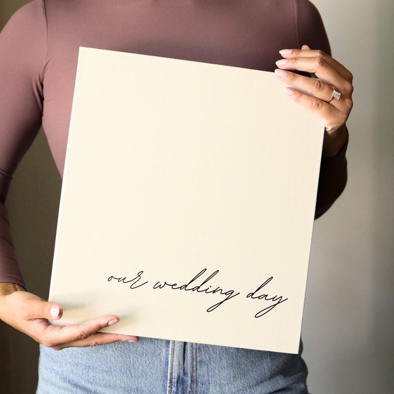 May include: A square, off-white canvas with the phrase "our wedding day" in elegant black script. The canvas is held by a person wearing a mauve long-sleeve shirt and blue jeans.