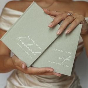 Peut inclure: Deux livres de v&oelig;ux de mariage vert sauge sont tenus. Le livre de gauche dit "Mes v&oelig;ux &agrave; mon mari le jour de notre mariage." Le livre de droite dit "Mes v&oelig;ux &agrave; ma femme le jour de notre mariage."