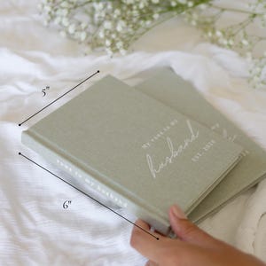 Peut inclure: Deux carnets de v&oelig;ux vert sauge, chacun mesurant 15 cm x 13 cm, avec le texte "My vows to my husband" et "EST. 2025" en &eacute;criture blanche. Les carnets sont sur une surface blanche, avec une branche de fleurs blanches en arri&egrave;re-plan.
