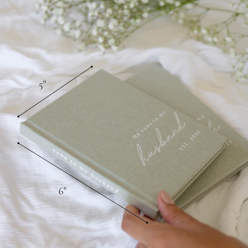 Peut inclure: Deux carnets de v&oelig;ux vert sauge, chacun mesurant 15 cm x 13 cm, avec le texte "My vows to my husband" et "EST. 2025" en &eacute;criture blanche. Les carnets sont sur une surface blanche, avec une branche de fleurs blanches en arri&egrave;re-plan.