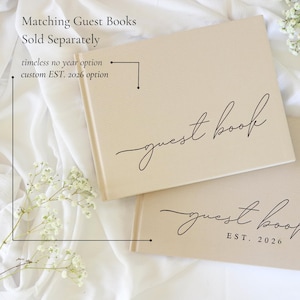 Peut inclure: Deux livres d'or beiges avec l'inscription "guest book" en &eacute;criture &eacute;l&eacute;gante. Un livre porte la mention "EST. 2026" en dessous. Le texte "Matching Guest Books Sold Separately" est en haut. Les livres sont sur un tissu blanc.