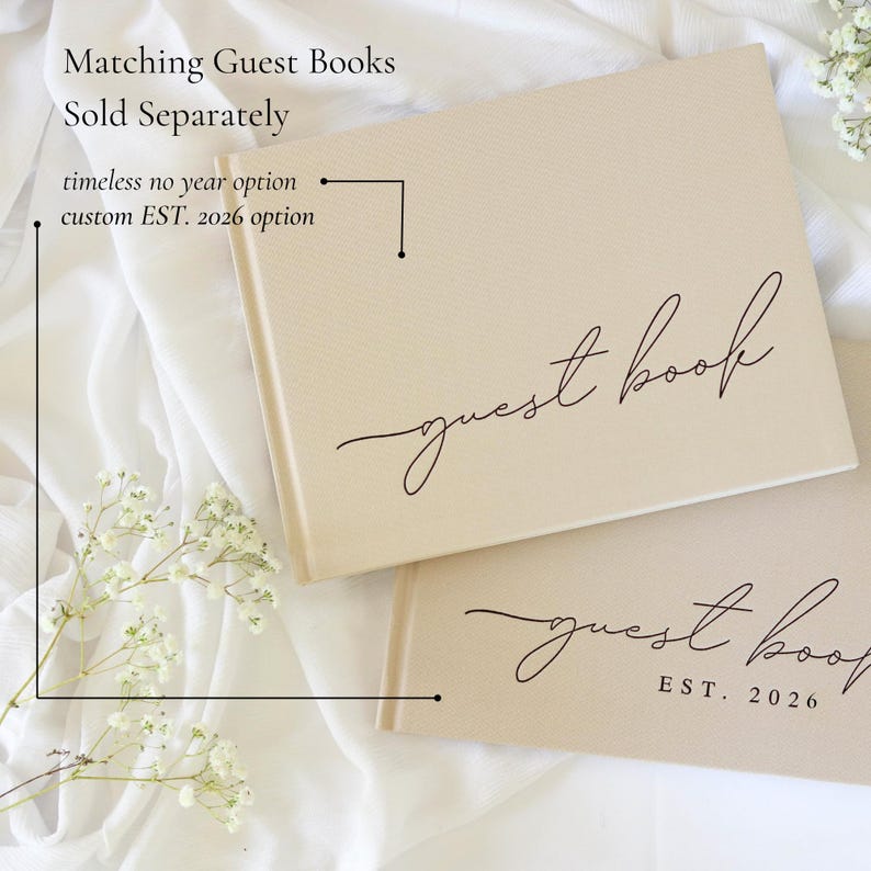 Peut inclure: Deux livres d'or beiges avec l'inscription "guest book" en &eacute;criture &eacute;l&eacute;gante. Un livre porte la mention "EST. 2026" en dessous. Le texte "Matching Guest Books Sold Separately" est en haut. Les livres sont sur un tissu blanc.