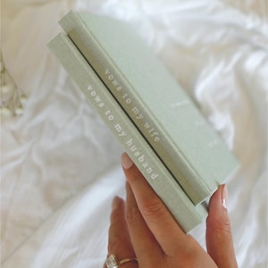 Peut inclure: Deux livres de v&oelig;ux vert sauge sont empil&eacute;s, le livre du dessus l&eacute;g&egrave;rement inclin&eacute;. Le texte "Vows to my wife" et "Vows to my husband" est imprim&eacute; en blanc sur les tranches. Les livres sont tenus par une main avec une bague.