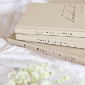 Peut inclure: Trois livres beiges empil&eacute;s, avec du texte noir sur les tranches. Le livre du dessus indique "My vows to my husband" et "EST. 2020". Le livre du milieu dit "vows to my husband" et le livre du bas dit "vows to my wife". Le livre du bas indique "Guest Book | EST. 2026".