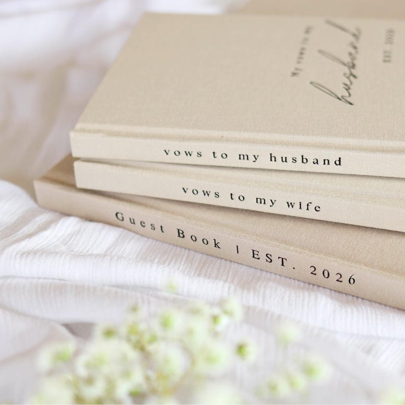 Peut inclure: Trois livres beiges empil&eacute;s, avec du texte noir sur les tranches. Le livre du dessus indique "My vows to my husband" et "EST. 2020". Le livre du milieu dit "vows to my husband" et le livre du bas dit "vows to my wife". Le livre du bas indique "Guest Book | EST. 2026".