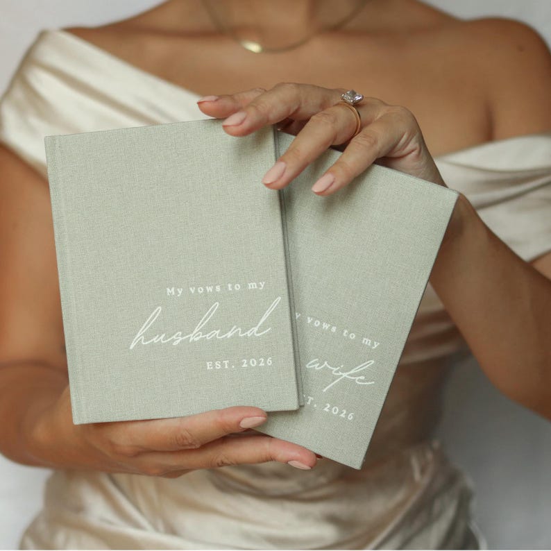 Peut inclure: Deux livres de v&oelig;ux de mariage vert sauge sont tenus devant une personne. Le livre de gauche dit "Mes v&oelig;ux &agrave; mon mari" avec "EST. 2026" en dessous. Le livre de droite dit "Mes v&oelig;ux &agrave; ma femme" avec "EST. 2026" en dessous.
