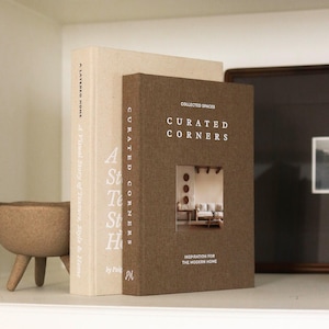May include: Two hardcover books on a shelf. The book on the left is beige with the title "A LAYERED HOME" and "A Visual Story of Texture, Style & Home". The book on the right is brown with the title "CURATED CORNERS" and "INSPIRATION FOR THE MODERN HOME".