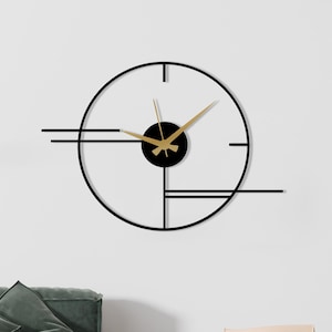 May include: A modern, minimalist wall clock with a black frame and gold hands. The clock features a circular design with geometric lines and a black center. The clock is mounted on a white wall above a green sofa.