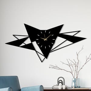 May include: A black geometric wall clock with gold hands and markers. The clock is mounted on a white wall. The clock's design features a starburst shape with triangular elements. The room also includes a blue armchair and a wooden cabinet.