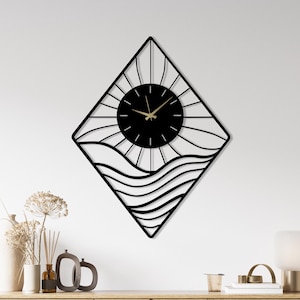 May include: Black metal wall clock with a diamond-shaped frame. The clock face is black with gold hands, and the design includes sun rays and wavy lines. The clock is mounted on a white wall above a wooden cabinet.