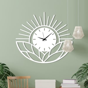 May include: A white, sun-shaped wall clock with black hands and a minimalist design. The clock is mounted on a light green wall, above a wooden desk and chair. Two hanging pendant lights and potted plants complete the decor.