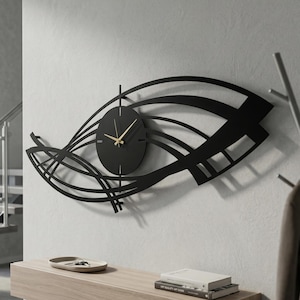 May include: A modern black wall clock with a unique, abstract design. The clock features a circular face with gold-colored hands, set against a backdrop of curved and linear black metal elements. The clock is mounted on a light gray wall.