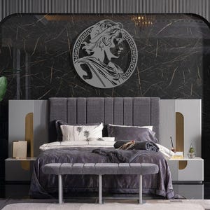 May include: A grey upholstered bed with a grey headboard and a grey bench at the foot of the bed. The headboard has a decorative design with vertical lines. The bed is decorated with a grey and white throw blanket and a grey pillow with a white design. A round metal wall decor with a Greek design is hanging on the wall behind the bed.