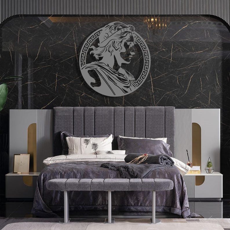 May include: A grey upholstered bed with a grey headboard and a grey bench at the foot of the bed. The headboard has a decorative design with vertical lines. The bed is decorated with a grey and white throw blanket and a grey pillow with a white design. A round metal wall decor with a Greek design is hanging on the wall behind the bed.