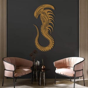 May include: A gold alien creature wall decal with a detailed design. The decal is on a dark gray wall with two pink armchairs and a glass table in front of it.