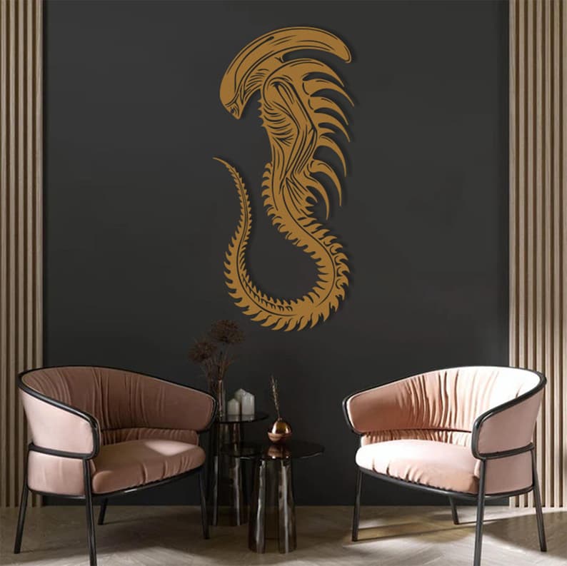 May include: A gold alien creature wall decal with a detailed design. The decal is on a dark gray wall with two pink armchairs and a glass table in front of it.
