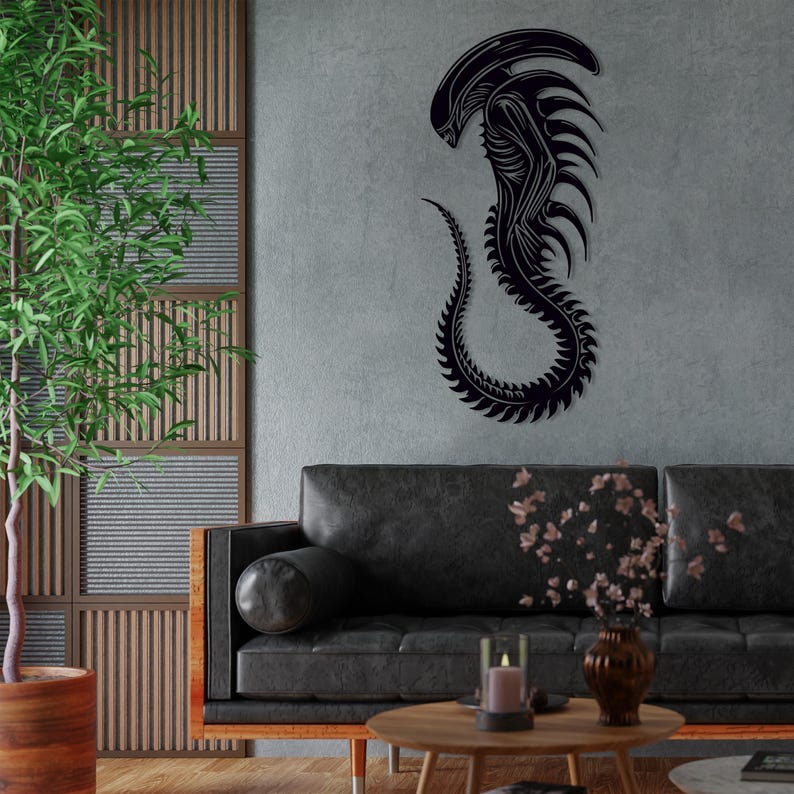 May include: Black metal wall art depicting a stylized alien creature with a long, segmented tail. The art is mounted on a gray wall in a living room setting with a black leather sofa and a coffee table.