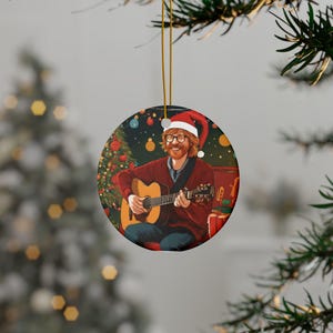 May include: Round Christmas ornament featuring a person playing a guitar and wearing a Santa hat. The ornament has a gold string for hanging. The background includes a Christmas tree and presents, creating a festive holiday scene.
