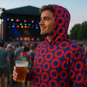 May include: A person wearing a blue hoodie with a red circle pattern, holding a clear plastic cup of beer at an outdoor concert. The background features a stage with colorful lights and a crowd of people.