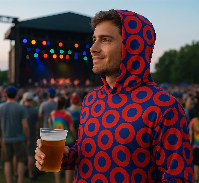 May include: A person wearing a blue hoodie with a red circle pattern, holding a clear plastic cup of beer at an outdoor concert. The background features a stage with colorful lights and a crowd of people.