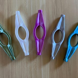May include: Five different coloured bookmark holders. The holders are made of plastic and have a unique shape. The colours are green, white, pink, grey, and blue.
