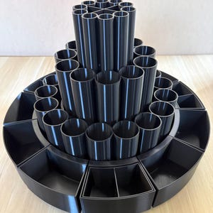 May include: A tiered, black organizer with multiple compartments. The top tier features cylindrical holders, while the lower tiers have open compartments. The organizer is designed to hold various items, such as makeup brushes or pens.