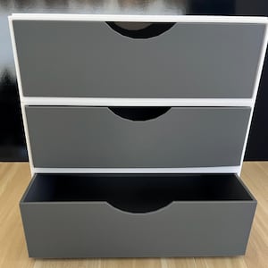 May include: A three-drawer storage unit with a white frame and gray drawers. The drawers have a curved handle design. The bottom drawer is open, revealing a dark interior. The unit is on a light wood surface.