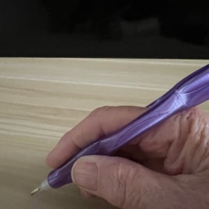 May include: A close-up of a hand holding a purple diamond painting pen with a metal tip. The pen is being used to place a diamond on a canvas.
