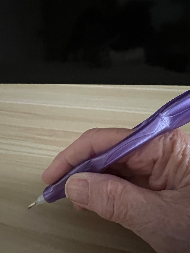May include: A close-up of a hand holding a purple diamond painting pen with a metal tip. The pen is being used to place a diamond on a canvas.
