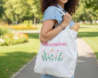 May include: White canvas tote bag with the name "Alexandra" in pink script and embroidered flowers. The bag is carried by a person in a denim dress and contains a loaf of bread. The background is a park with green trees and a walking path.