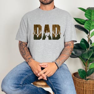 May include: A gray t-shirt with a brown and green graphic of the word "DAD" with a deer silhouette on each side.