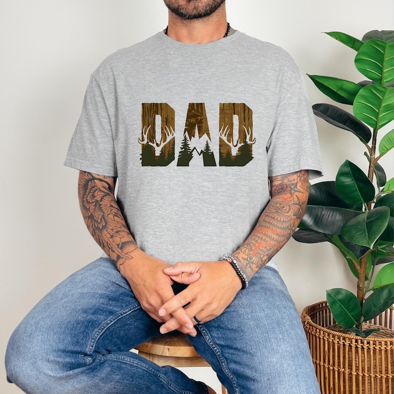 May include: A gray t-shirt with a brown and green graphic of the word "DAD" with a deer silhouette on each side.