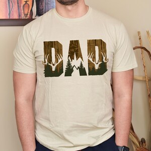 May include: A beige t-shirt with a graphic print of the word "DAD" in brown and green. The graphic features a mountain range, trees, and deer antlers.