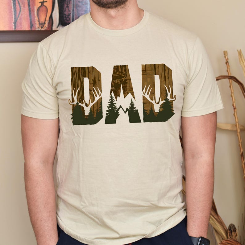 May include: A beige t-shirt with a graphic print of the word "DAD" in brown and green. The graphic features a mountain range, trees, and deer antlers.