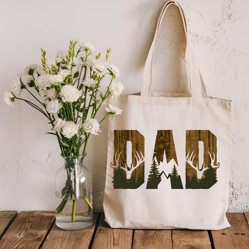 May include: A white canvas tote bag with the word "DAD" printed in brown on the front. The letters are surrounded by a wood grain pattern and images of deer antlers, trees, and mountains.
